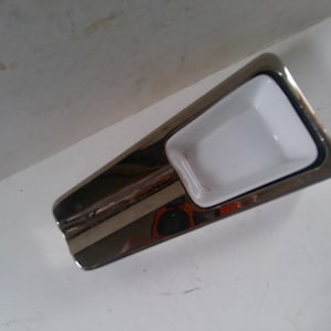 May include: A silver and white ashtray with a rectangular shape and a white ceramic insert. The ashtray has a smooth, polished surface.