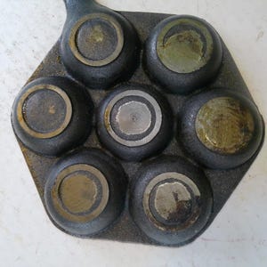 Może przedstawiać: Żeliwna patelnia z siedmioma okrągłymi wgłębieniami, każde z mosiężnym pierścieniem. Patelnia jest ciemnoszara z uchwytem wystającym z jednej strony. Wnętrze każdego wgłębienia ma nieco inne metaliczne wykończenie.