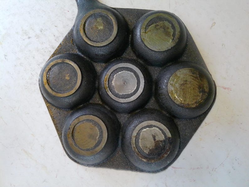 Może przedstawiać: Żeliwna patelnia z siedmioma okrągłymi wgłębieniami, każde z mosiężnym pierścieniem. Patelnia jest ciemnoszara z uchwytem wystającym z jednej strony. Wnętrze każdego wgłębienia ma nieco inne metaliczne wykończenie.