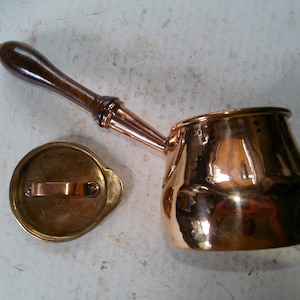 May include: A copper-colored, small pot with a wooden handle and a matching lid. The pot has a rounded body and a spout. The lid is round with a copper handle. The handle is dark brown.