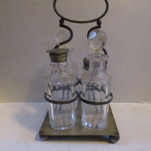 May include: A vintage condiment set featuring two clear glass bottles with faceted stoppers and metal lids, held in a metal stand. The stand has a handle and a rectangular base. The bottles are approximately 4 inches tall.