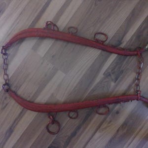 May include: Red painted horse harness with metal rings and chains. The harness is curved and has a decorative brass ball on each end. The harness is lying on a wooden surface.