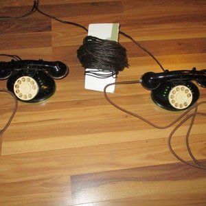 May include: Two vintage black rotary dial telephones with cream-colored dials and brown cords. The phones are connected to a bundle of black wire, suggesting a communication setup. The phones are on a wooden surface.