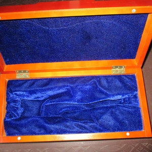 Pu&ograve; includere: Scatola di legno aperta con un esterno marrone rossiccio intenso e un interno in velluto blu vibrante. La scatola &egrave; rettangolare con cerniere in ottone e una fodera morbida e soffice, che suggerisce un astuccio per gioielli o altri oggetti di valore.