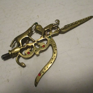 May include: A gold-colored metal ornament with a detailed design of animals and a long, pointed tail. The ornament has red and green gemstones embedded in it.