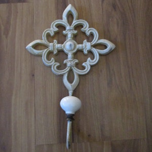 May include: Ornate, off-white metal wall hook with a fleur-de-lis design. Features a white ceramic knob and a curved hook. Decorative accent for entryways, bedrooms, or bathrooms. Measures approximately 7 inches in length.