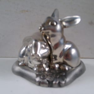 May include: A silver figurine of two rabbits, one sitting on top of the other. The rabbits are both facing forward and have their ears up. The figurine is on a base with a floral design.