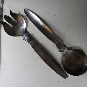 May include: A vintage salad serving set featuring a fork and spoon. The fork has three tines, and both pieces have dark wooden handles and ornate silver-toned metal detailing. The set is designed for serving salads.