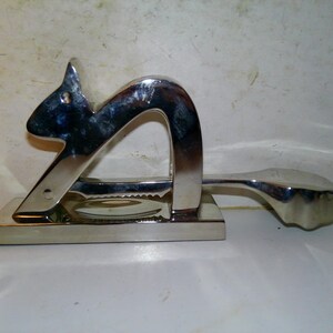 May include: A silver-colored letter opener in the shape of a squirrel. The opener has a rectangular base and a curved handle. The squirrel's head is at the top, and the tail forms the handle.
