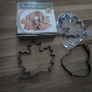 May include: A set of three metal pancake molds in the shape of maple leaves. The molds are packaged in a box with a picture of pancakes on it. The box says "Gourmet Pancake Molds".