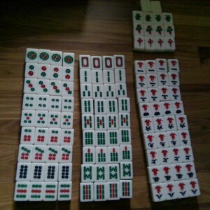 Pu&ograve; includere: Una collezione di tessere bianche da Mahjong disposte in tre colonne verticali e un piccolo gruppo. Le tessere presentano vari simboli incisi in verde e rosso, tra cui cerchi, bastoncini di bamb&ugrave;, caratteri cinesi e motivi floreali.