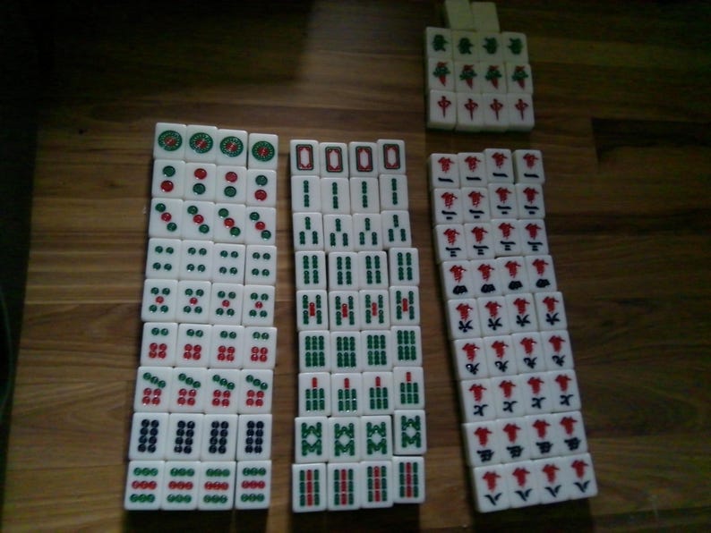 Pu&ograve; includere: Una collezione di tessere bianche da Mahjong disposte in tre colonne verticali e un piccolo gruppo. Le tessere presentano vari simboli incisi in verde e rosso, tra cui cerchi, bastoncini di bamb&ugrave;, caratteri cinesi e motivi floreali.