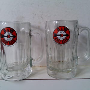 May include: Two clear glass A&W Root Beer mugs with handles. Each mug has a red and white circular logo with the words "A&W Ice Cold Root Beer" and "Trademark Reg. U.S. Pat. Off."