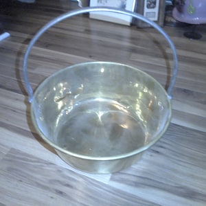 May include: A brass bucket with a silver handle. The bucket is empty and sitting on a wooden floor.