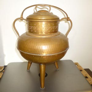 May include: A vintage copper pot with a lid and handle. The pot has a rounded body with a hammered texture and sits on three legs. The lid and upper section feature decorative floral patterns. The pot is a warm, golden color.