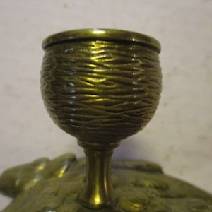 K&ouml;nnte beinhalten: Nahaufnahme eines messingfarbenen Dekorationsgegenstands. Der becherf&ouml;rmige obere Teil weist ein strukturiertes, gewebtes Design auf. Der schlanke Stiel f&uuml;hrt zu einem blattf&ouml;rmigen Sockel. Das Objekt hat eine verzierte, Vintage-&Auml;sthetik.