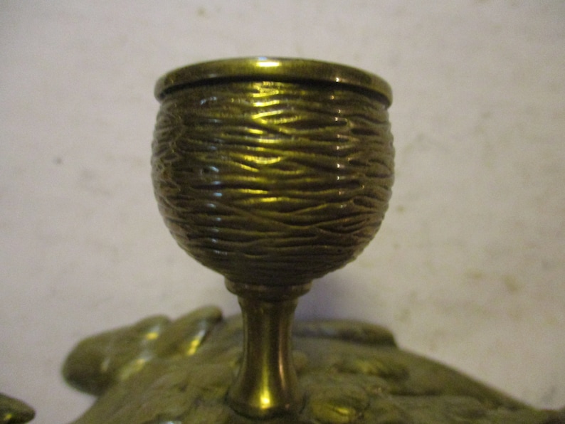 K&ouml;nnte beinhalten: Nahaufnahme eines messingfarbenen Dekorationsgegenstands. Der becherf&ouml;rmige obere Teil weist ein strukturiertes, gewebtes Design auf. Der schlanke Stiel f&uuml;hrt zu einem blattf&ouml;rmigen Sockel. Das Objekt hat eine verzierte, Vintage-&Auml;sthetik.