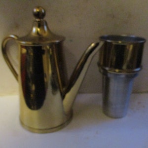May include: A gold-colored metal teapot with a lid and a matching gold-colored metal spout. The teapot is sitting on a white surface next to a silver-colored metal object.