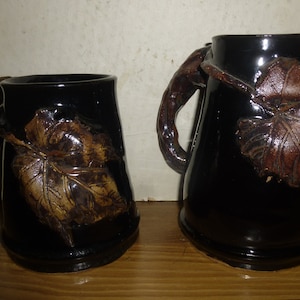 May include: Two black ceramic mugs with brown leaf designs. The mug on the right has a handle shaped like a vine.