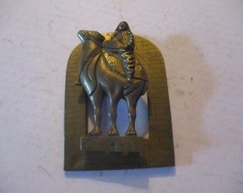 May include: A brass-toned metal sculpture of a camel with a rider, set against a brick-patterned arch. The camel is detailed with textured saddle and rider's clothing. The sculpture is mounted on a rectangular base.