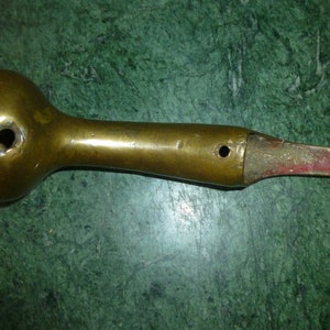 May include: A vintage brass tool with a bulbous head and a long, slender handle. The handle has a hole near the head and at the end. The tool shows signs of age and wear, with a reddish-brown section at the end.