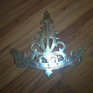 May include: A brass protractor with an ornate design. The protractor has a scale from 0 to 20 degrees.