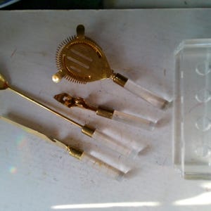May include: A collection of gold-toned bar tools with clear acrylic handles, including a strainer, spoon, and knife. A clear acrylic holder is also present, suggesting a bar or cocktail theme.