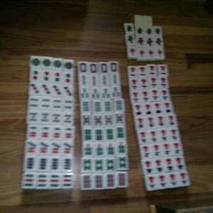 Pu&ograve; includere: Una collezione di tessere rettangolari bianche da mahjong disposte in tre colonne verticali e un piccolo gruppo. Ogni tessera presenta simboli, caratteri e motivi numerici incisi con inchiostro rosso, verde e nero, organizzati per seme.