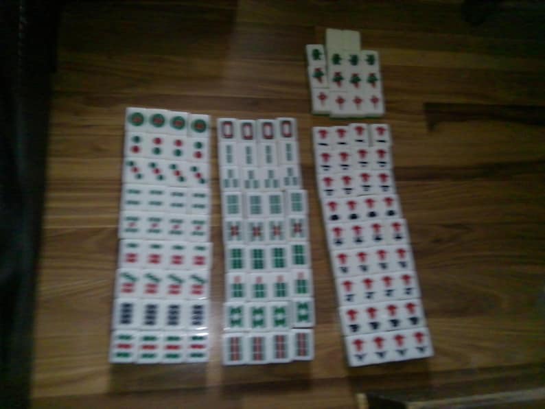 Pu&ograve; includere: Una collezione di tessere rettangolari bianche da mahjong disposte in tre colonne verticali e un piccolo gruppo. Ogni tessera presenta simboli, caratteri e motivi numerici incisi con inchiostro rosso, verde e nero, organizzati per seme.