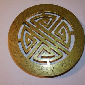 Puede incluir: Un salvamanteles redondo de color latón con un intrincado diseño calado. El centro presenta un patrón geométrico, rodeado por un borde floral. El salvamanteles se utiliza probablemente para proteger las superficies de los objetos calientes.