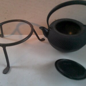 May include: A black cast iron teapot with a textured surface and a matching lid. The teapot is sitting on a black metal stand with three legs.