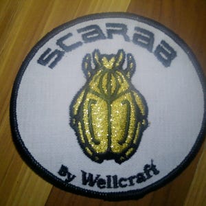May include: Circular patch with a white background, black border, and the word "SCARAB" in gray block letters. A gold, glittery scarab beetle is in the center. The text "By Wellcraft" is below the beetle.