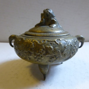 May include: A small, antique brass incense burner with a lid featuring a decorative animal figure. The burner has three legs and intricate floral designs.