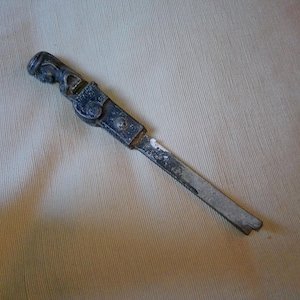 May include: An antique metal nutcracker with a detailed handle and a silver-colored blade. The nutcracker is dark gray with signs of wear and tear, suggesting age and use. It rests on a beige fabric surface.