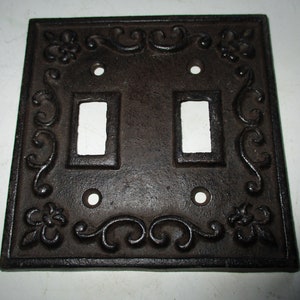 May include: A square, dark brown cast iron double light switch plate. The plate has two rectangular openings for the switches and decorative floral and scrollwork around the edges. There are also screw holes for mounting.