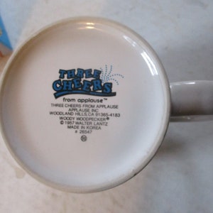 Puede incluir: Taza de cer&aacute;mica blanca con un logotipo azul y blanco que dice "Three Cheers from Applause" con un dise&ntilde;o de fuegos artificiales azules. El logotipo tambi&eacute;n incluye el texto "Three Cheers from Applause", "Applause Inc", "Woodland Hills, CA 91365-4183", "Woody Woodpecker", "&copy; 1957 Walter Lantz", "Made in Korea", y "# 26547".