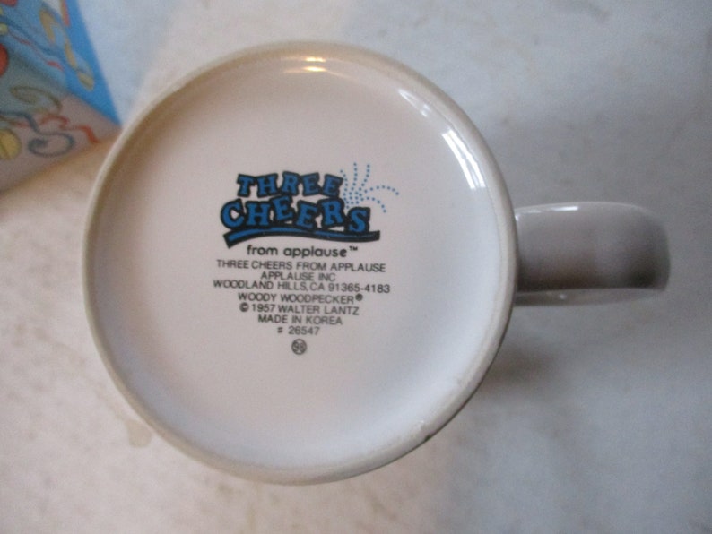 Puede incluir: Taza de cer&aacute;mica blanca con un logotipo azul y blanco que dice "Three Cheers from Applause" con un dise&ntilde;o de fuegos artificiales azules. El logotipo tambi&eacute;n incluye el texto "Three Cheers from Applause", "Applause Inc", "Woodland Hills, CA 91365-4183", "Woody Woodpecker", "&copy; 1957 Walter Lantz", "Made in Korea", y "# 26547".