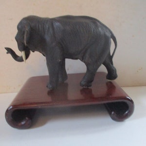 May include: A black elephant figurine with ivory tusks standing on a dark wood base. The elephant is facing left and has a detailed, textured surface.