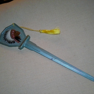 May include: A light blue letter opener with a shield-shaped handle featuring a Native American chief design. A yellow tassel hangs from the handle. The blade is long and pointed.