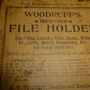 May include: Antique file holder with the text "WOODRUFF'S IMPROVED FILE HOLDER" printed on aged paper. The text details the holder's purpose for filing documents like letters and bills. The frame is made of wood.