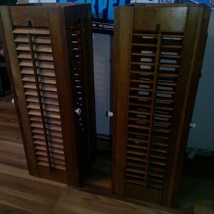 May include: Two tall, rectangular wooden shutters with a medium brown finish. Each shutter features horizontal slats arranged in a fixed, louvered pattern within a solid wood frame. Small white knobs are attached to the side of each shutter.
