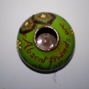 May include: A green ceramic trinket dish with brown floral designs and the text "To Absent Friends" in brown lettering.