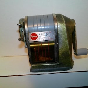 May include: Vintage green and gray Premier portable pencil sharpener. The sharpener has a hand crank and a label that reads "Premier Portable" with the Apso products inc. logo. The sharpener is made of metal and has a rectangular shape.