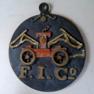 May include: A round, dark gray metal plaque with a decorative design. The plaque features a red and gold cart with the letters "F.I.Co" in gold. The top has a loop for hanging.