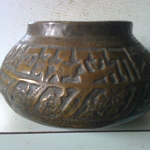 May include: A large, round, copper pot with intricate embossed designs. The pot has a wide mouth and a rounded bottom. The designs include floral motifs and geometric patterns.