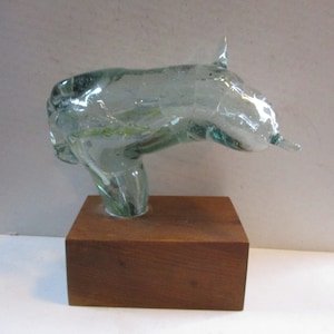 May include: A clear glass sculpture of a bull's head, mounted on a rectangular wooden base. The bull's head is detailed with horns and a snout, and the glass has a slightly green tint. The wooden base is a warm brown color.