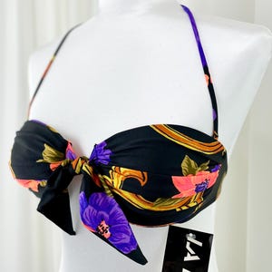 May include: Black bikini top with a floral pattern in pink, purple, and gold. The top has a bow in the center and thin purple straps. The product is displayed on a white mannequin.
