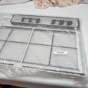 May include: A rectangular white plastic air conditioner filter frame with a black mesh grid interior, encased in clear bubble wrap. The frame features a small white rectangular label with a barcode and text printed on the surface of the mesh.