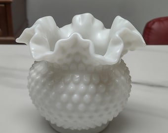 May include: A white hobnail glass vase with a ruffled rim. The vase has a round body covered in small, raised bumps. The ruffled rim adds a decorative touch to the classic design. This vase is perfect for displaying flowers or as a decorative accent.
