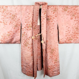 May include: A pale pink kimono featuring an all-over pattern of delicate floral motifs in darker pink and metallic gold tones. The garment has wide sleeves, a structured collar, and a small decorative cord tie at the front centre closure.